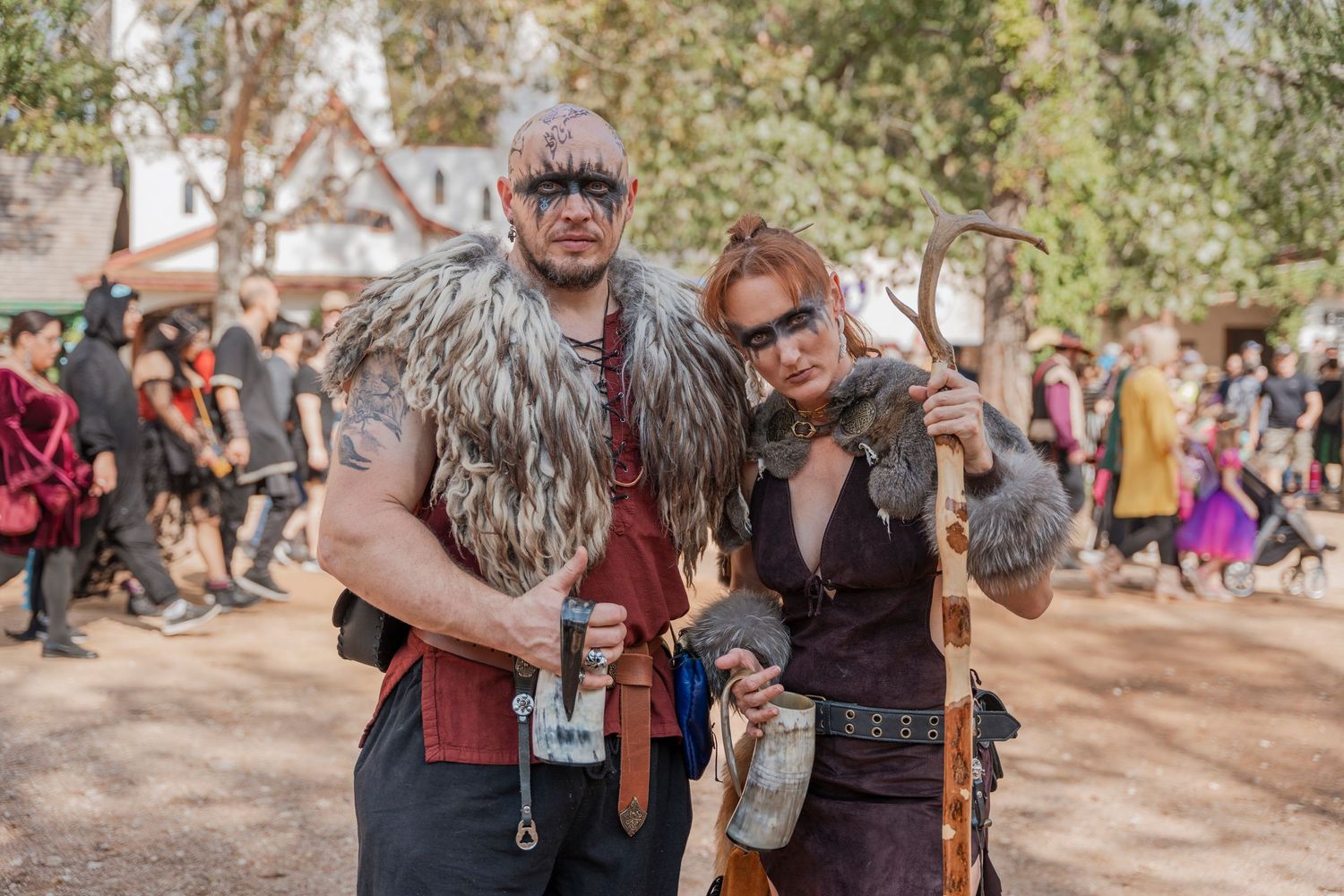 Two costumed festival attendees pose together wearing furs and medieval-style garments at a Renaissance fair.
