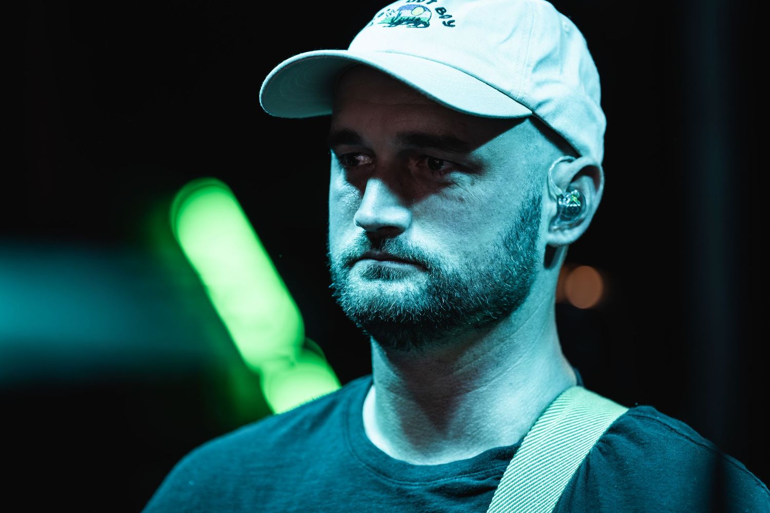 Musician in baseball cap performs under moody blue and green stage lighting.
