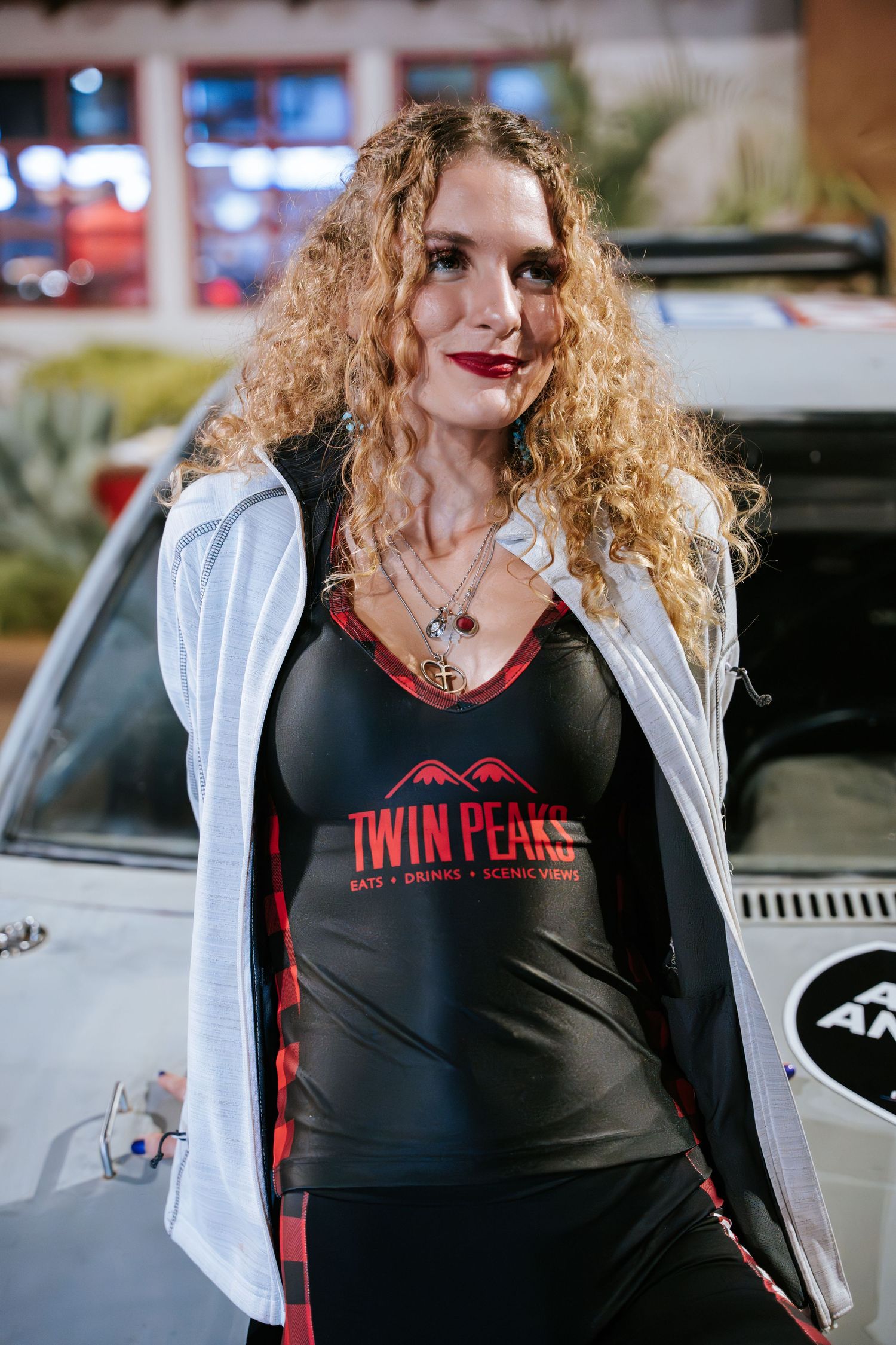 Woman in Twin Peaks branded tank top and white jacket at car show.
