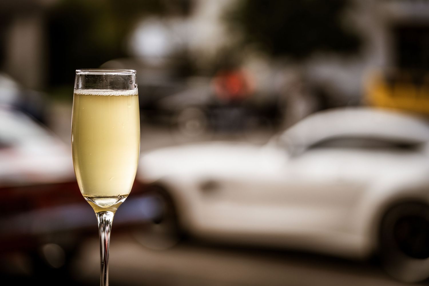 A champagne flute filled with bubbly beverage with luxury cars blurred in the background.