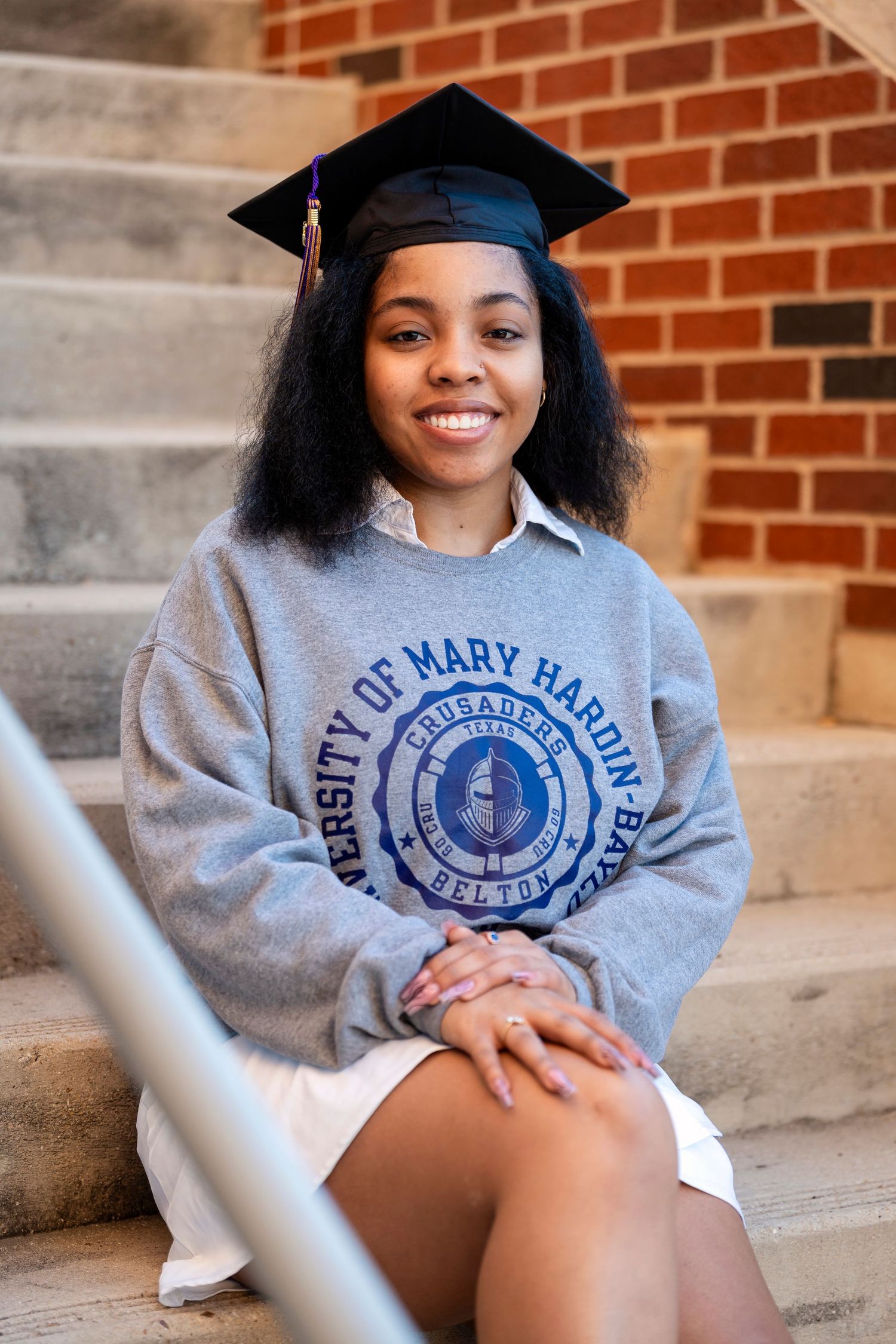 A graduation portrait on brick steps featuring academic regalia and university branded sweatshirt.