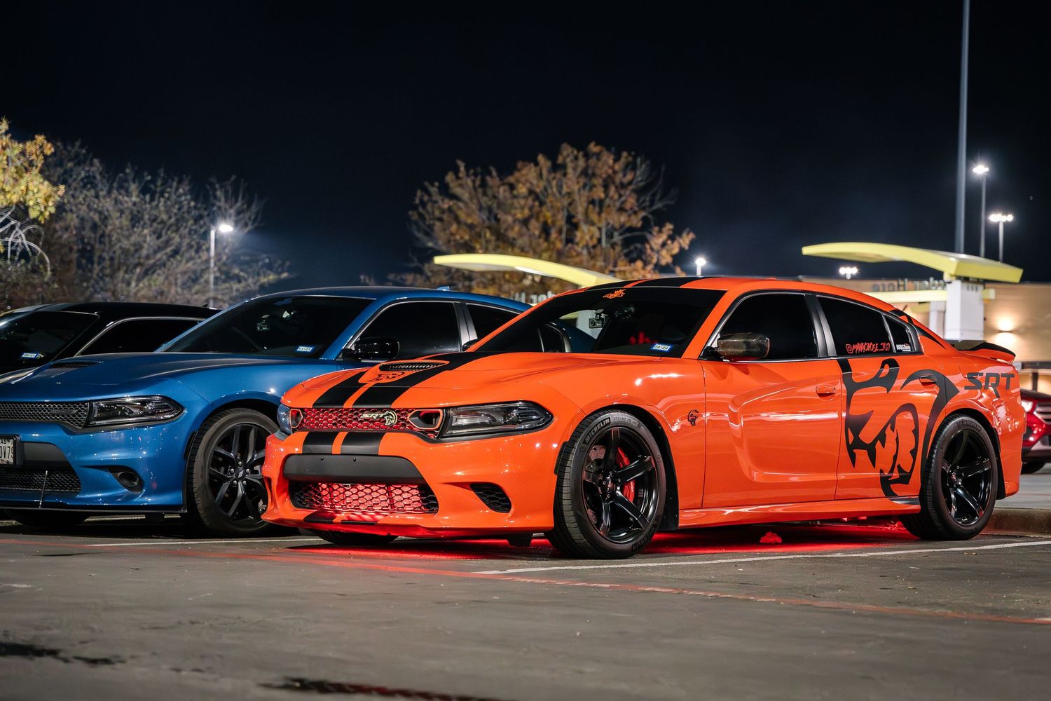 Two high-performance Dodge Chargers, one blue and one orange, parked under streetlights at night.