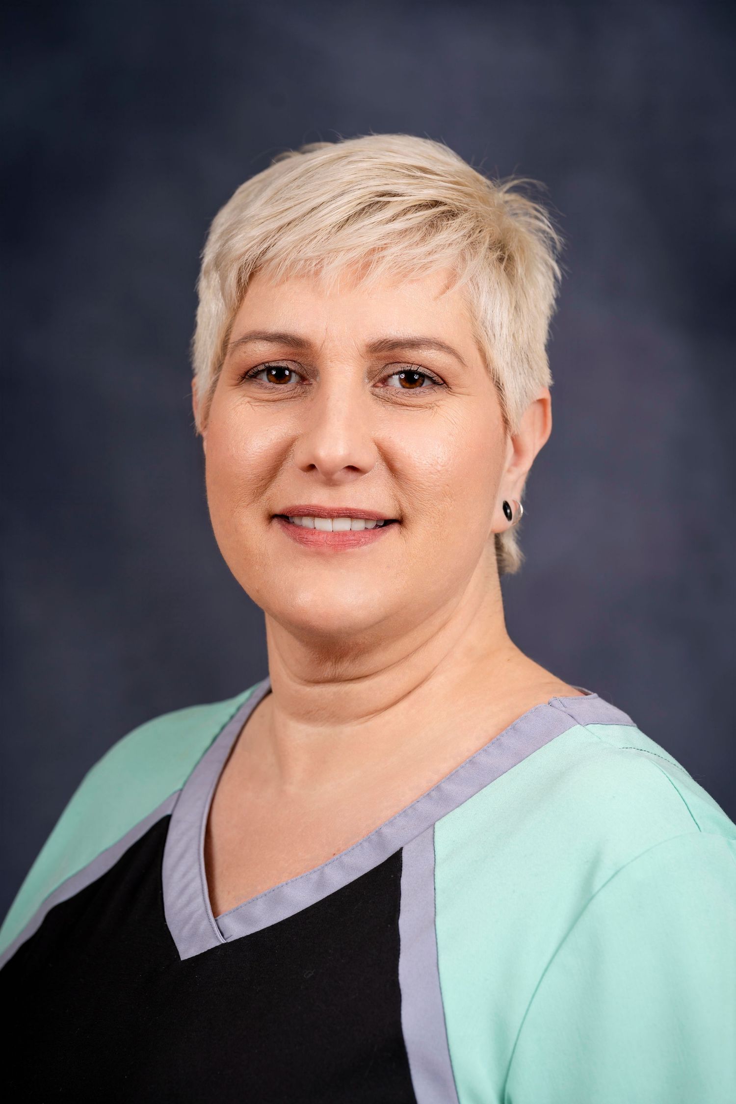 Professional headshot of a person with short blonde hair wearing mint green and gray medical attire.