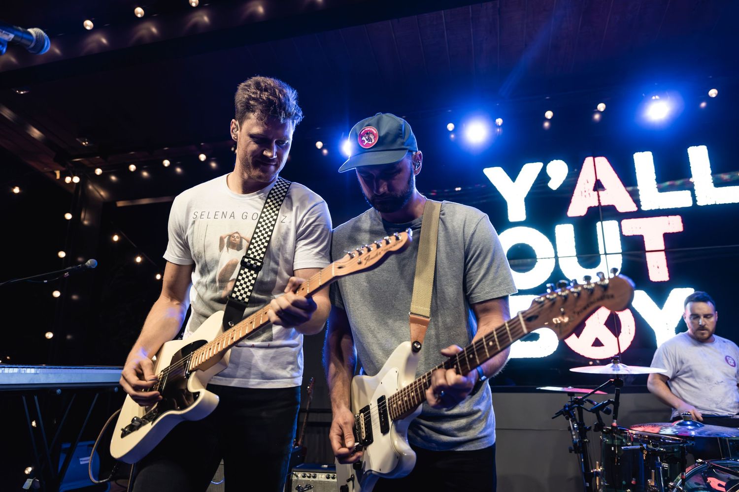 Musicians perform with guitars on stage with bright blue lighting and 'Y'ALL OUT' text display in the background.