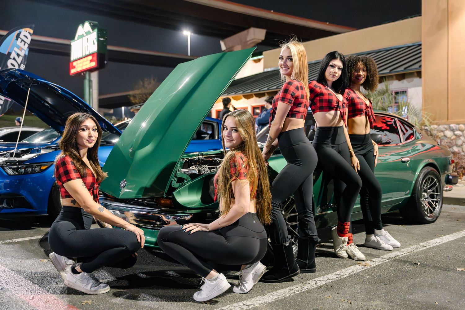 Group gathered around classic green car with open hood in nighttime parking lot.