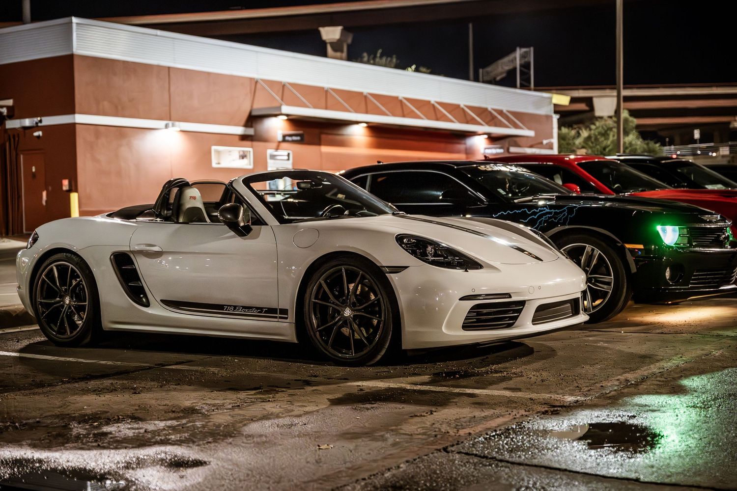White Porsche Cayman sports car with custom wheels parked in a wet parking lot at night.