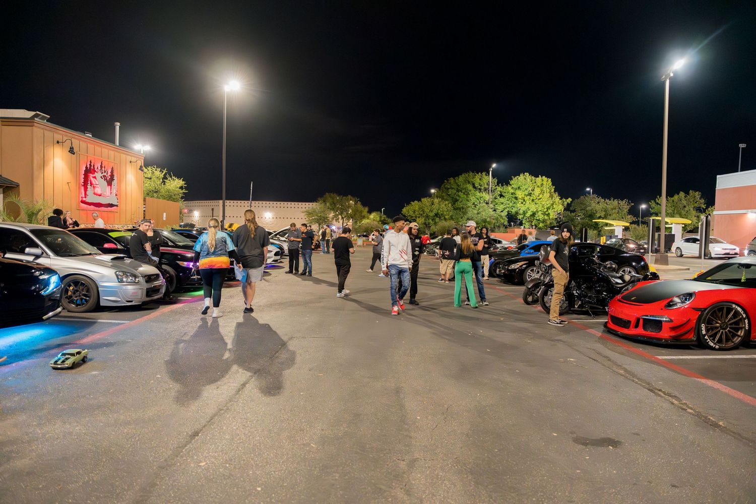 A nighttime car meet showing various modified vehicles in a parking lot.