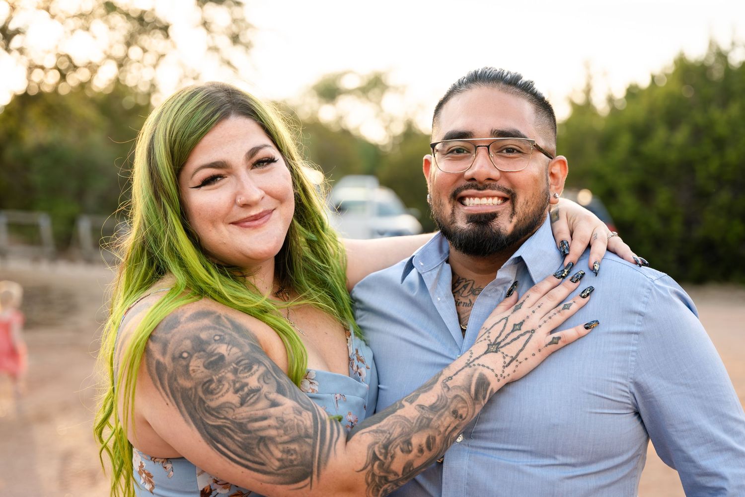 A couple showing off engagement ring with visible tattoos and green hair outdoors at sunset.