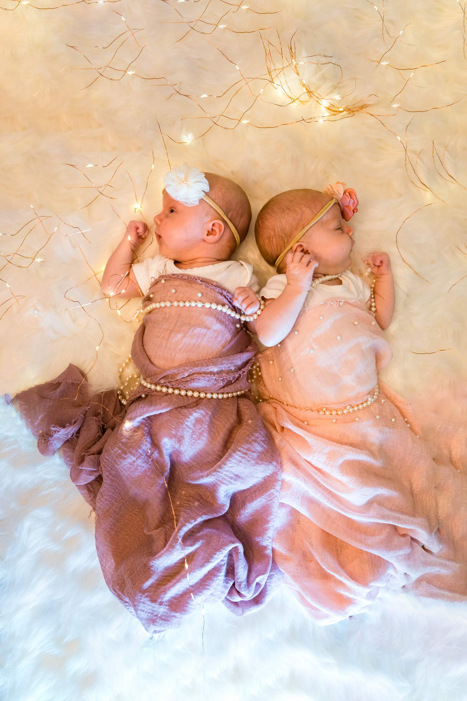 Two babies in pink and purple dresses lying on white fur with string lights overhead.
