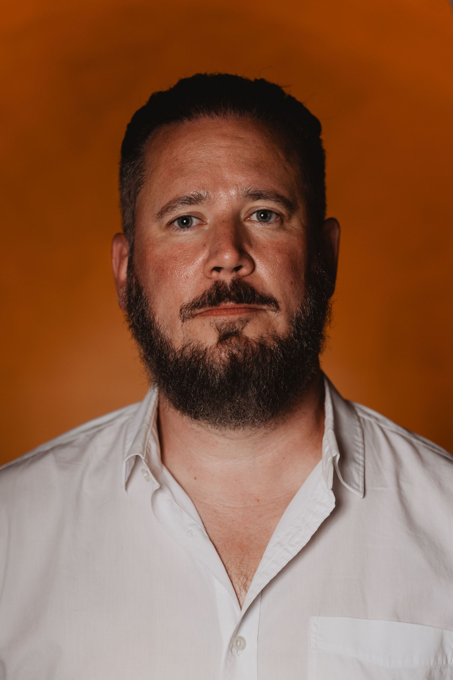 Portrait of a bearded man in white dress shirt against vibrant orange background.