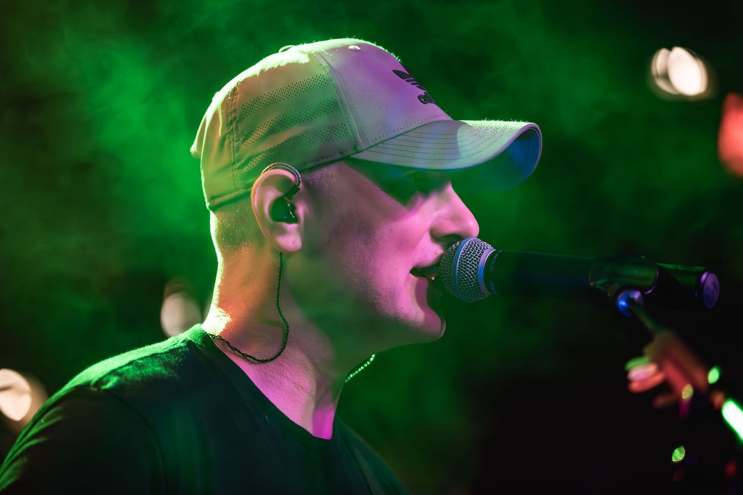 Performer in baseball cap singing into microphone with green stage lighting.