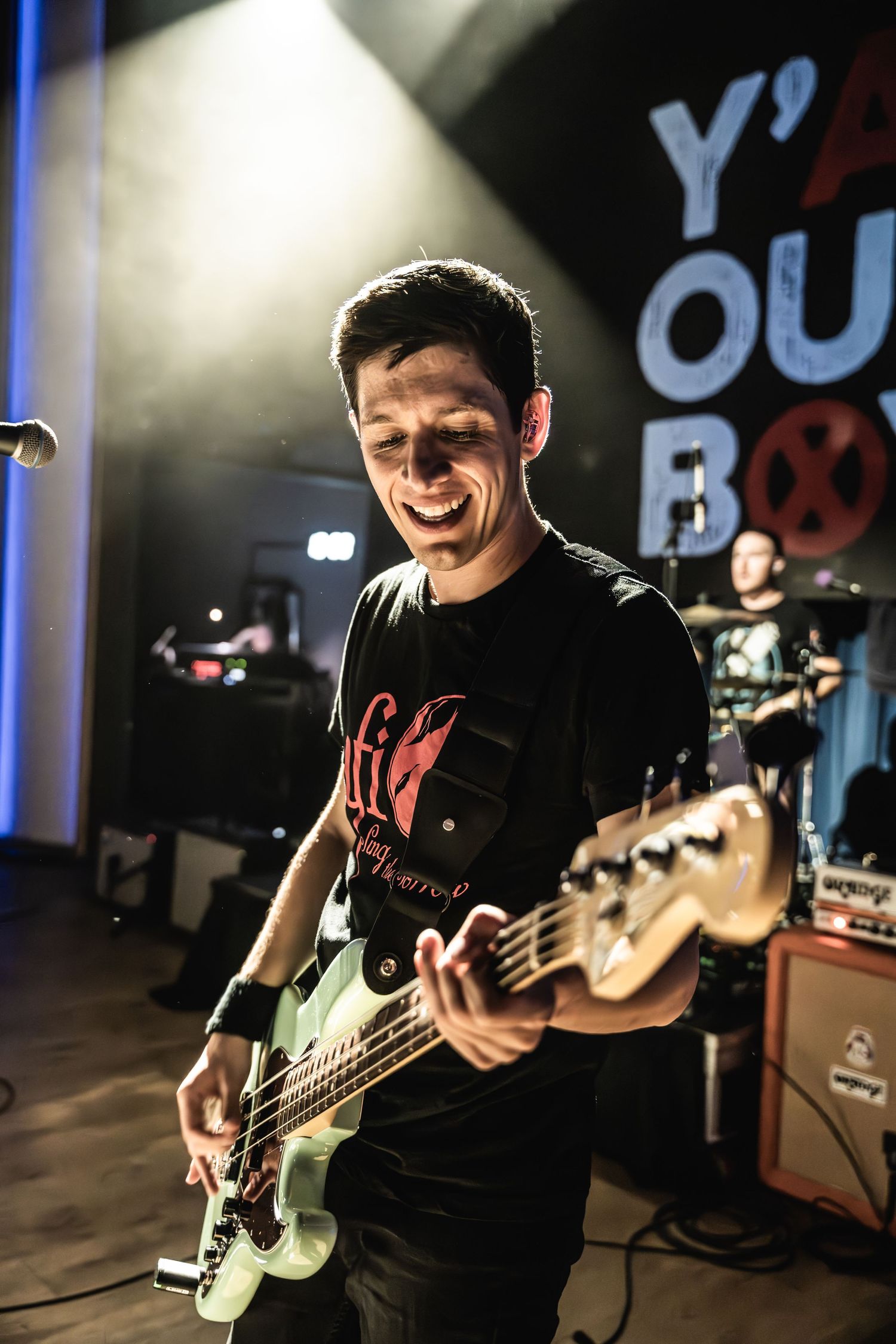 Bass guitarist performing enthusiastically on stage with 'Y' signage visible in background.