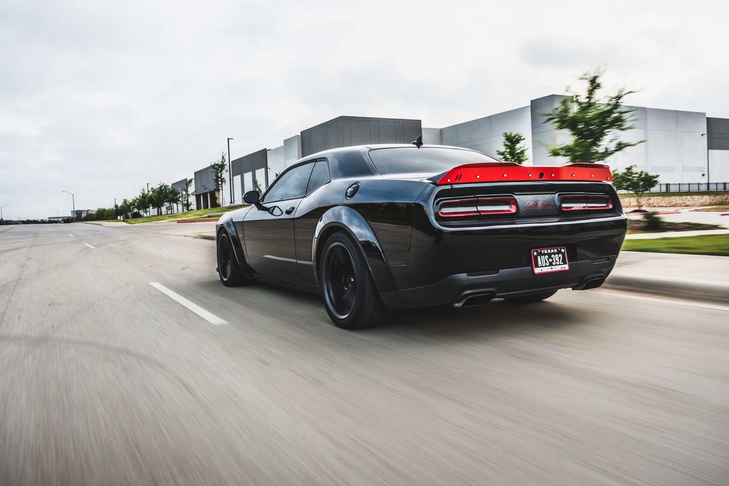 Rear view of a modified black Dodge Challenger with custom wheels driving on an industrial street.