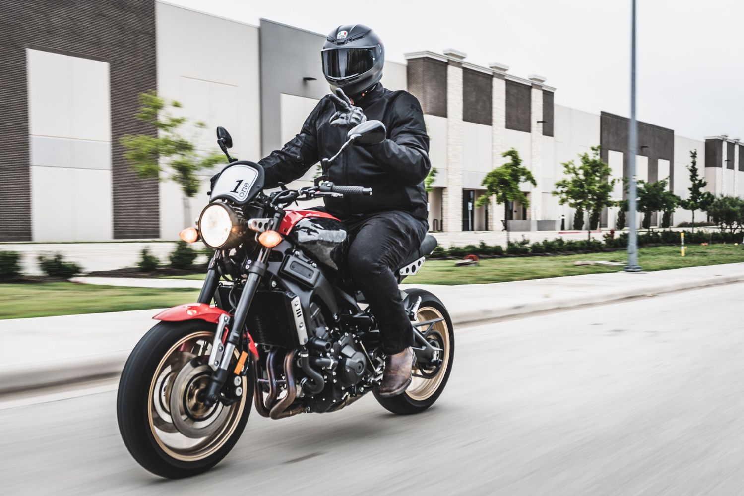 Motorcyclist in black gear rides a modern sport bike on an urban street with industrial buildings in background.