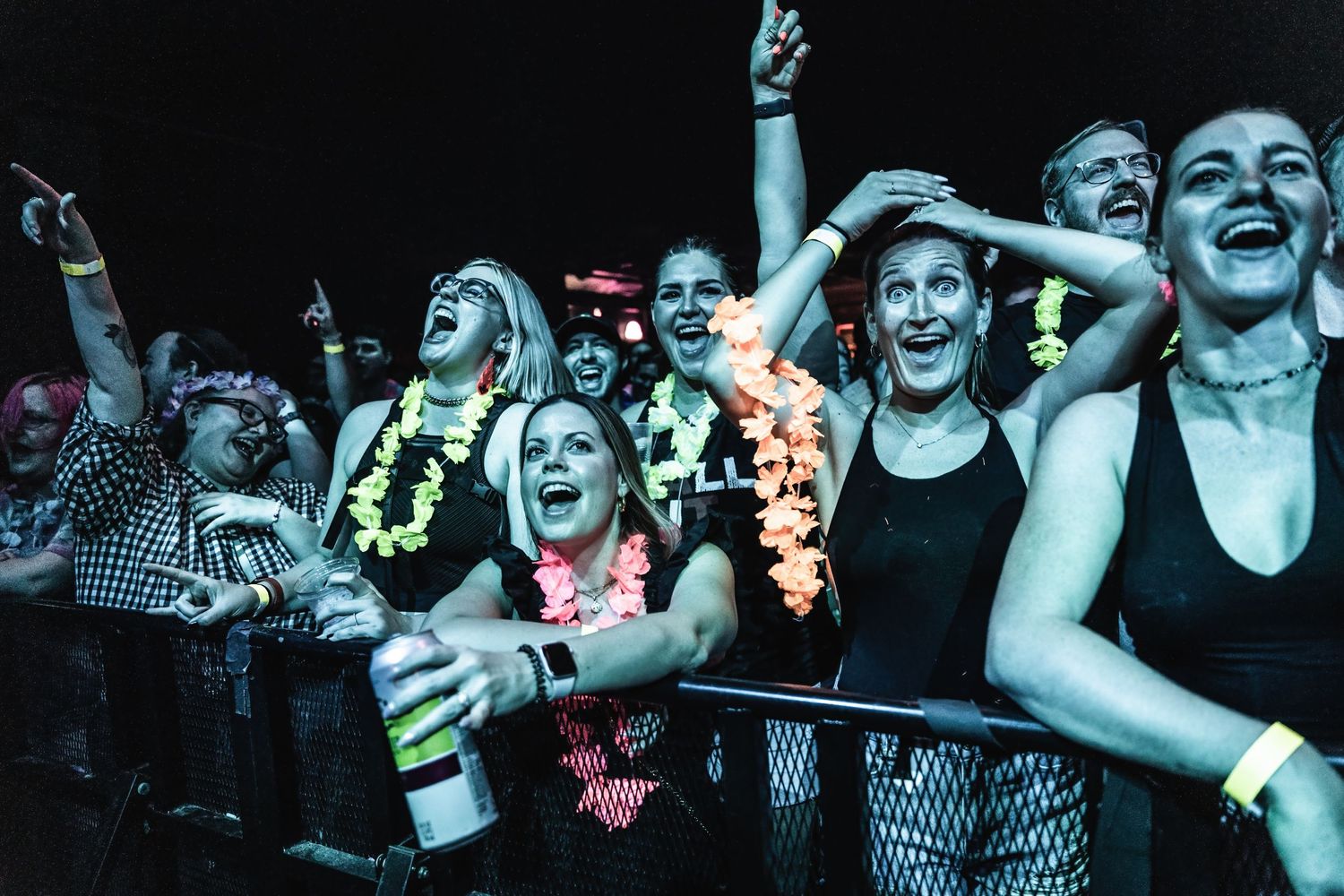 Energetic fans with flower leis cheer and raise hands at concert barrier under dramatic lighting.