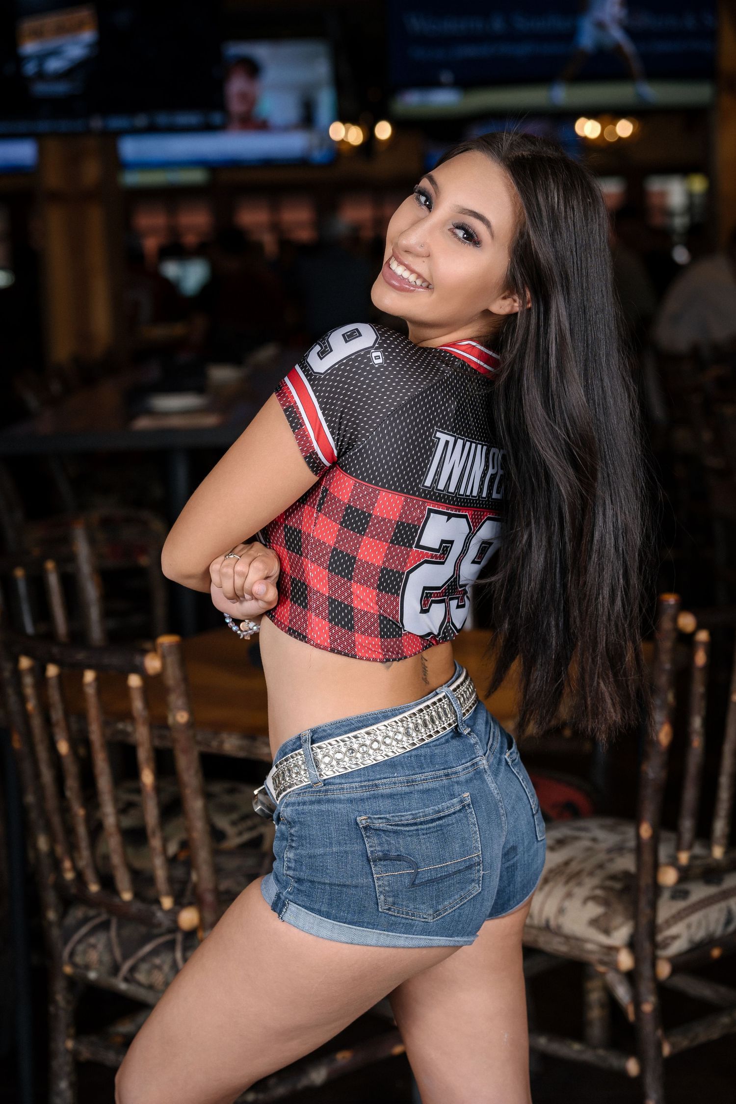 Restaurant staff member in plaid Twin Peaks crop top and denim shorts poses near bar seating.