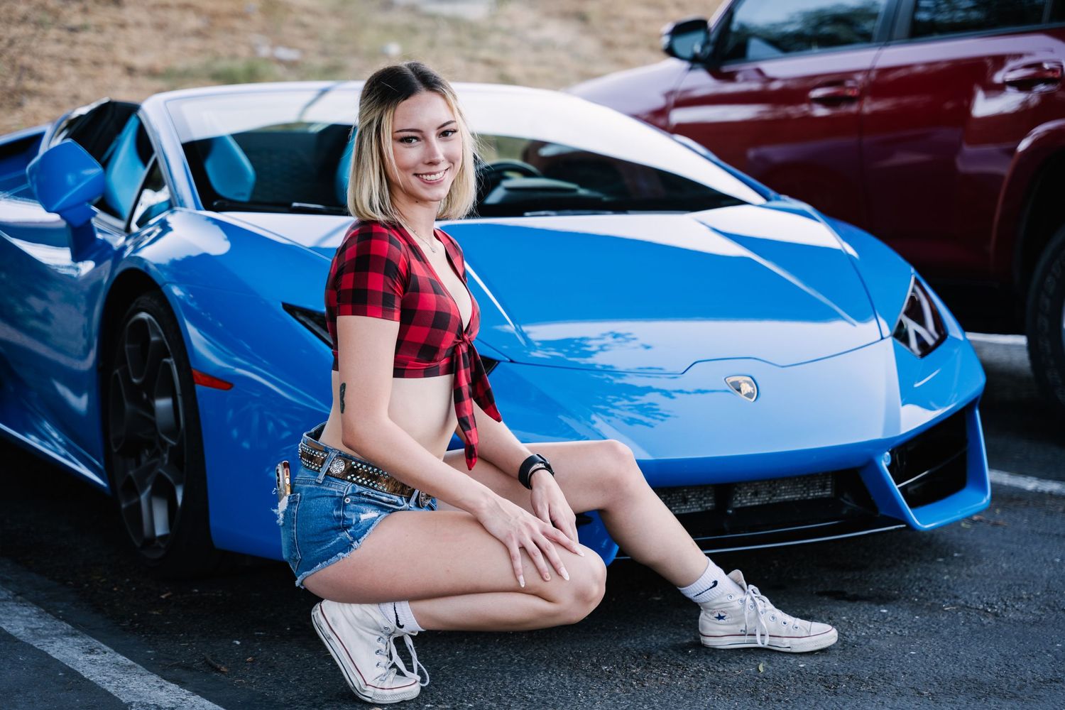 Model in red plaid shirt poses next to blue Lamborghini in parking lot.