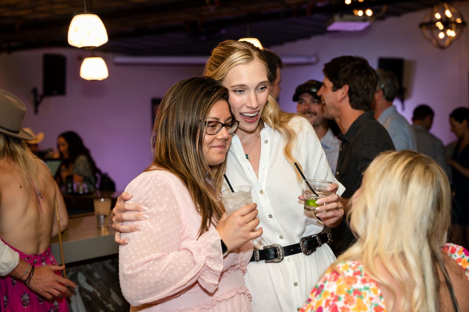 Friends sharing laughs and drinks at an indoor party with purple mood lighting.
