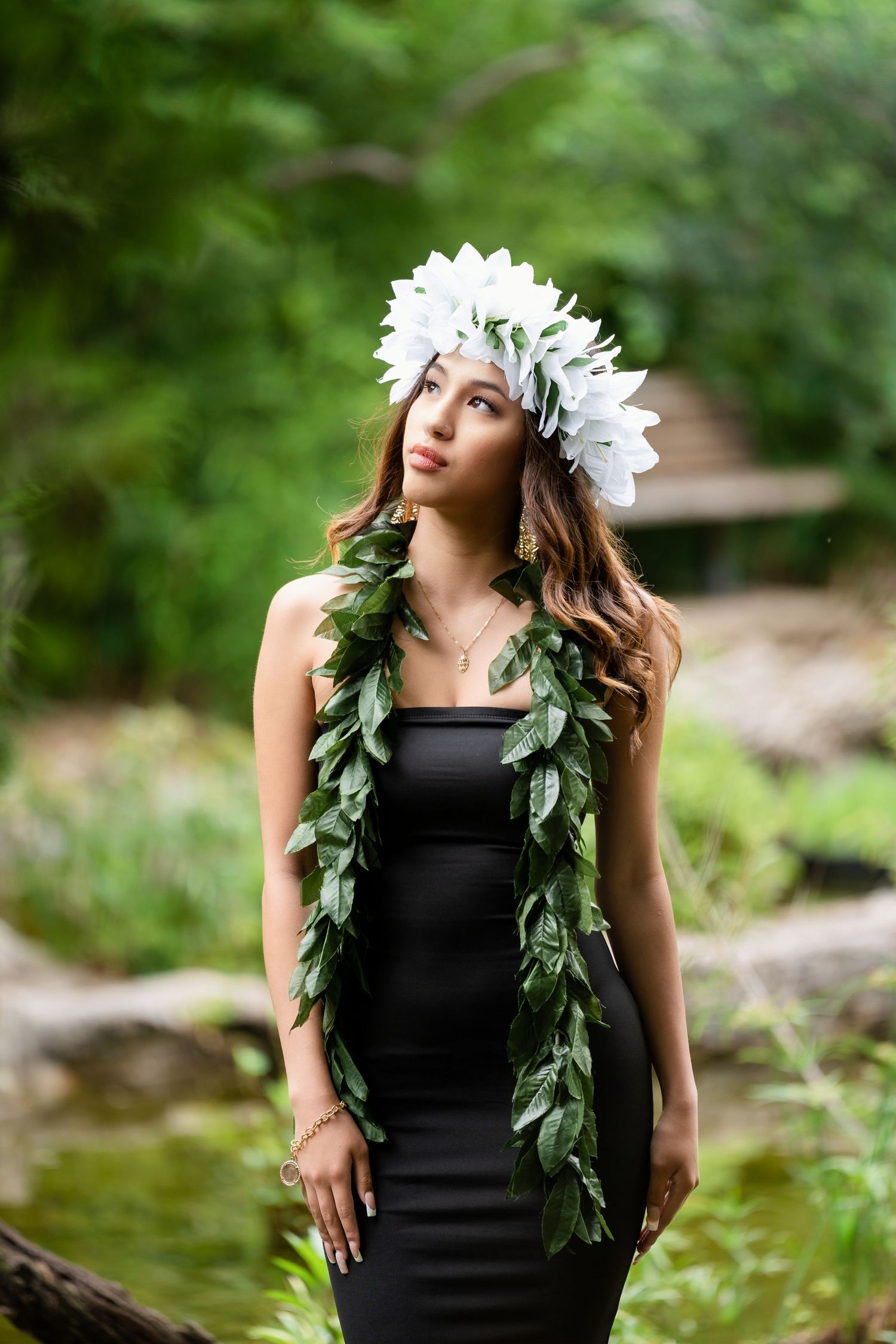 Model in black dress with leafy garland and white flower crown poses in garden setting.