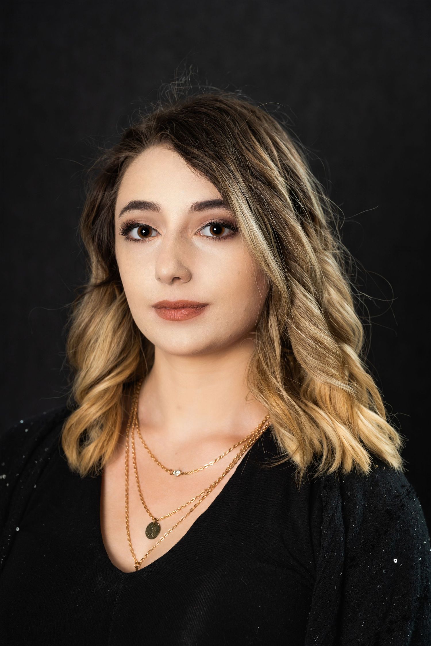 Studio portrait of woman wearing black top with layered necklaces against dark backdrop.