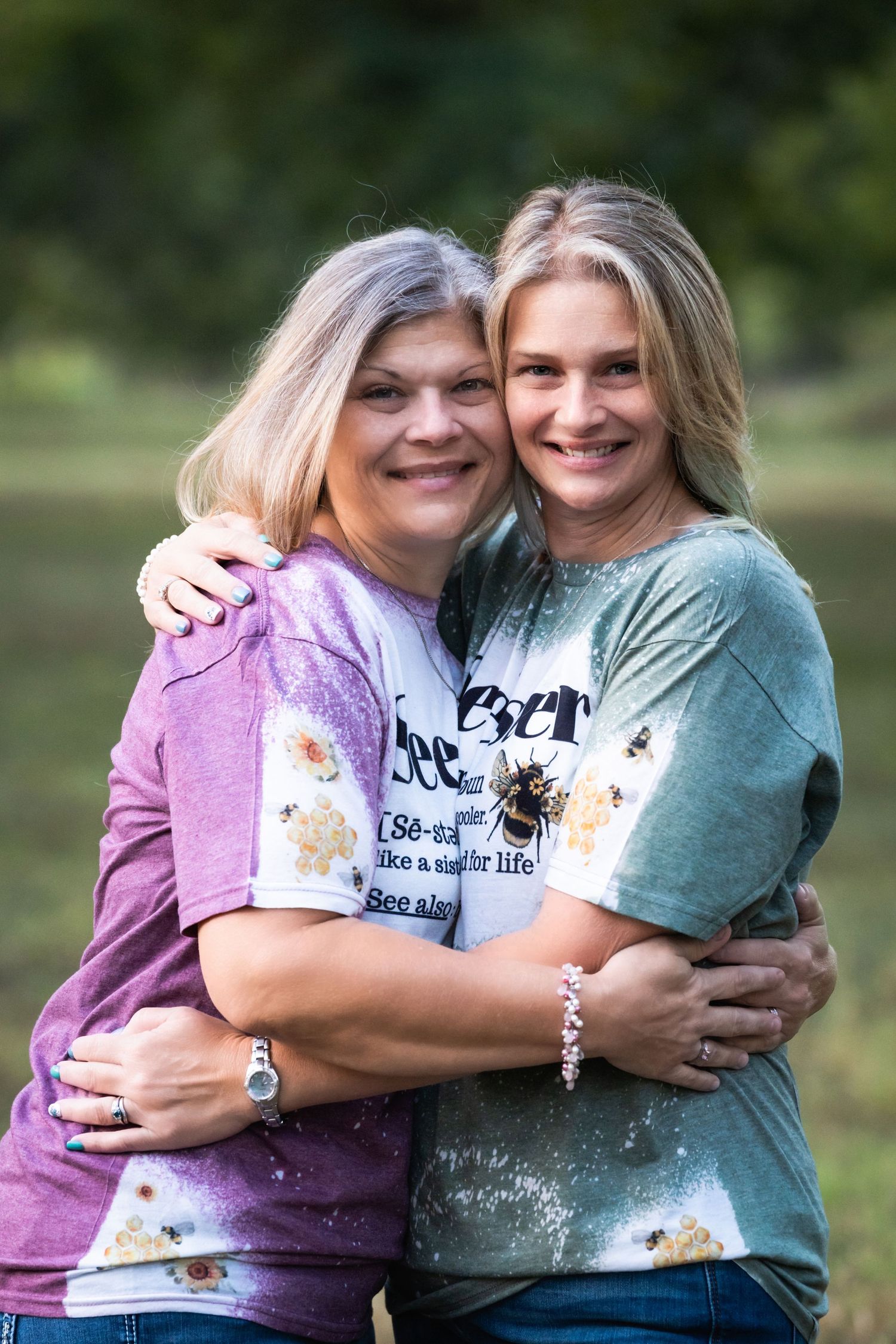 Two people wearing colorful t-shirts embrace while smiling outdoors in a natural setting.