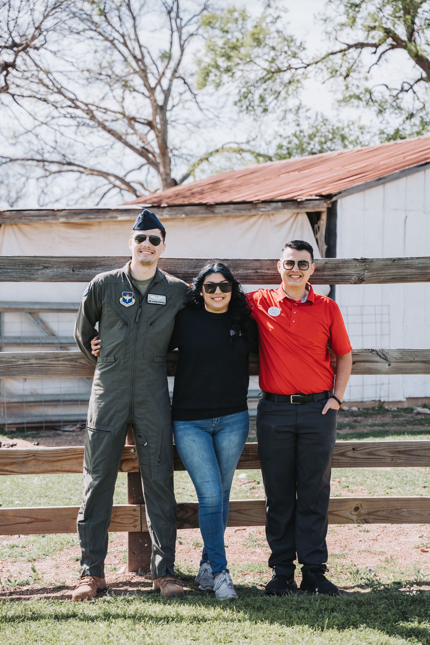 Group photo of three people standing by a wooden fence in casual outdoor attire.