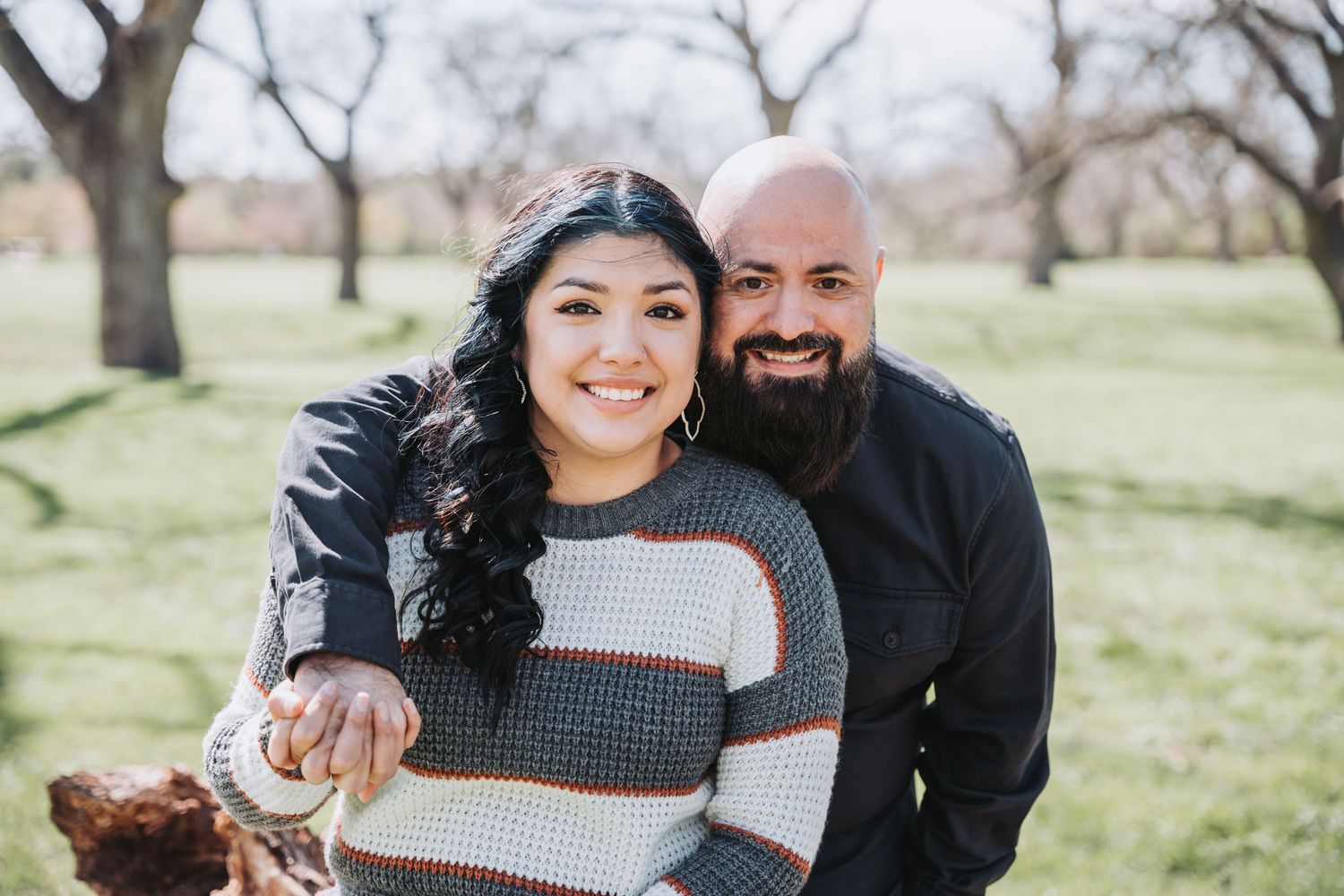 Happy couple poses together outdoors with bare trees and grass in background.