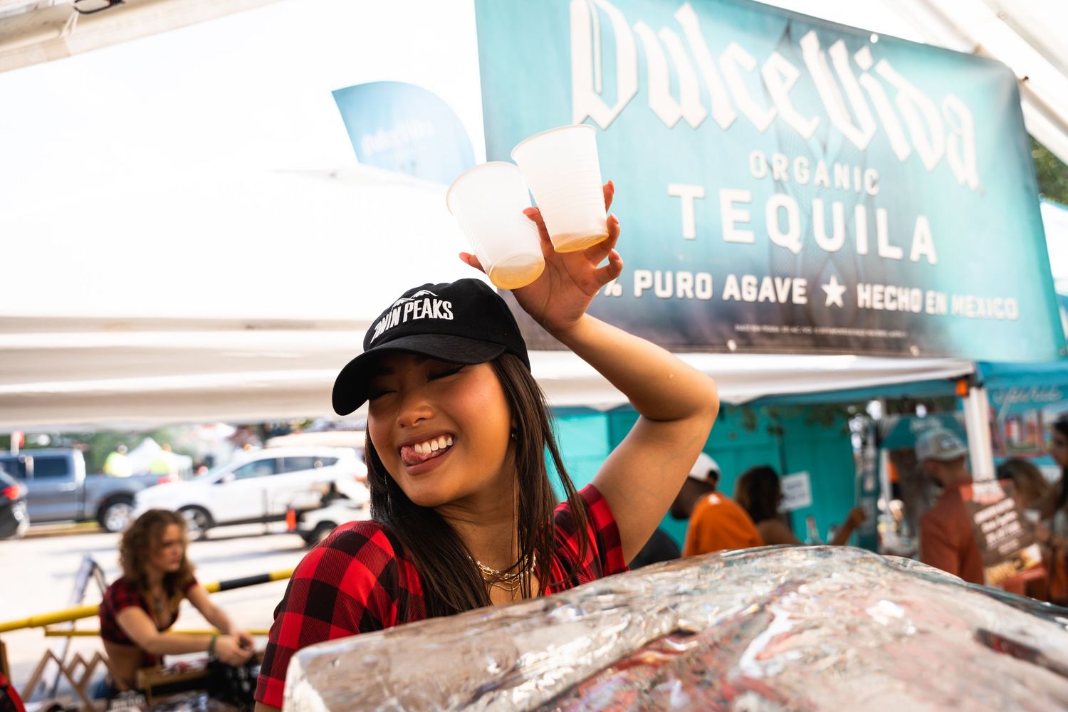 Beer festival attendee in red plaid shirt and baseball cap smiling at outdoor tequila tent.