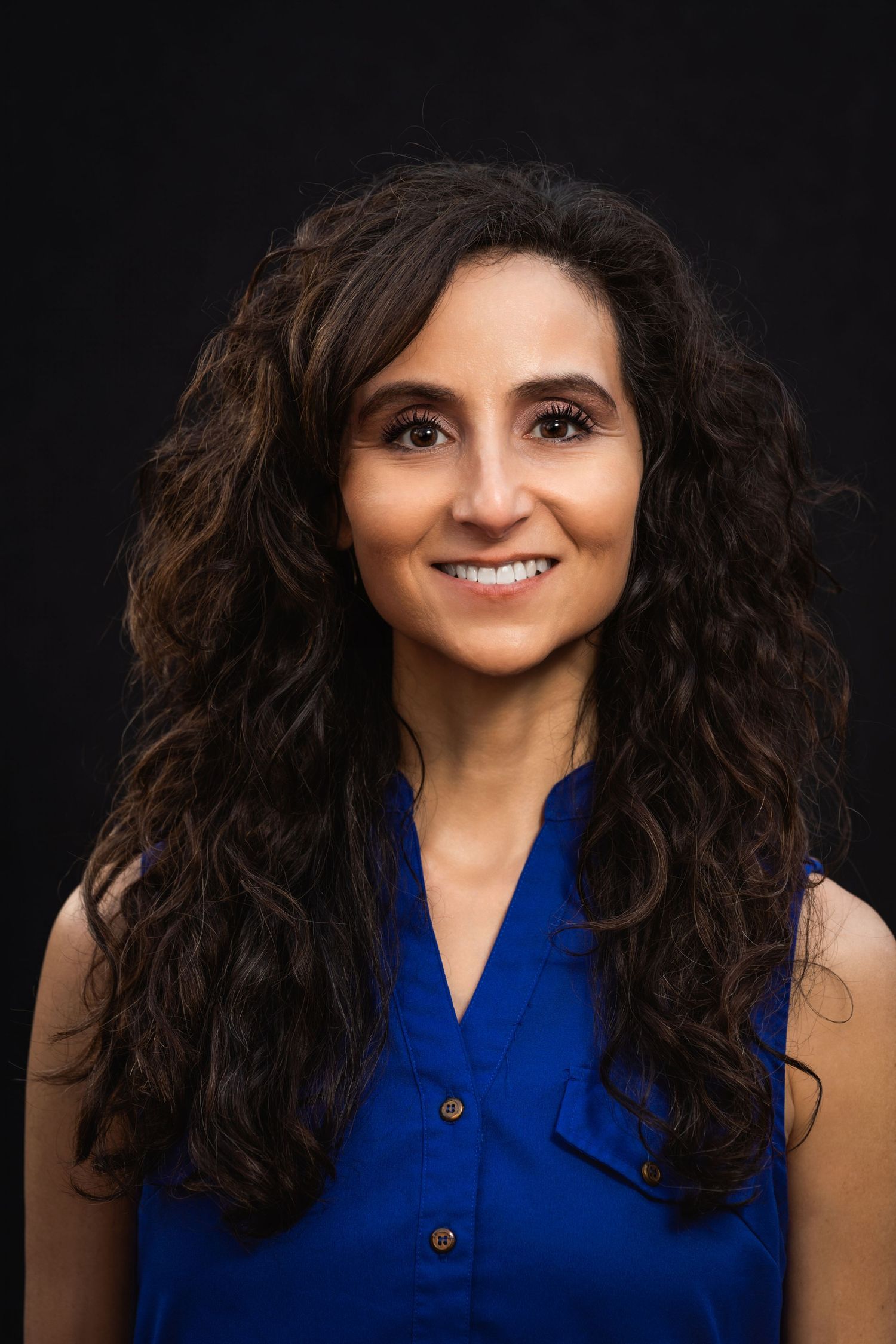 Professional headshot of person with curly dark hair wearing blue sleeveless top against black background.