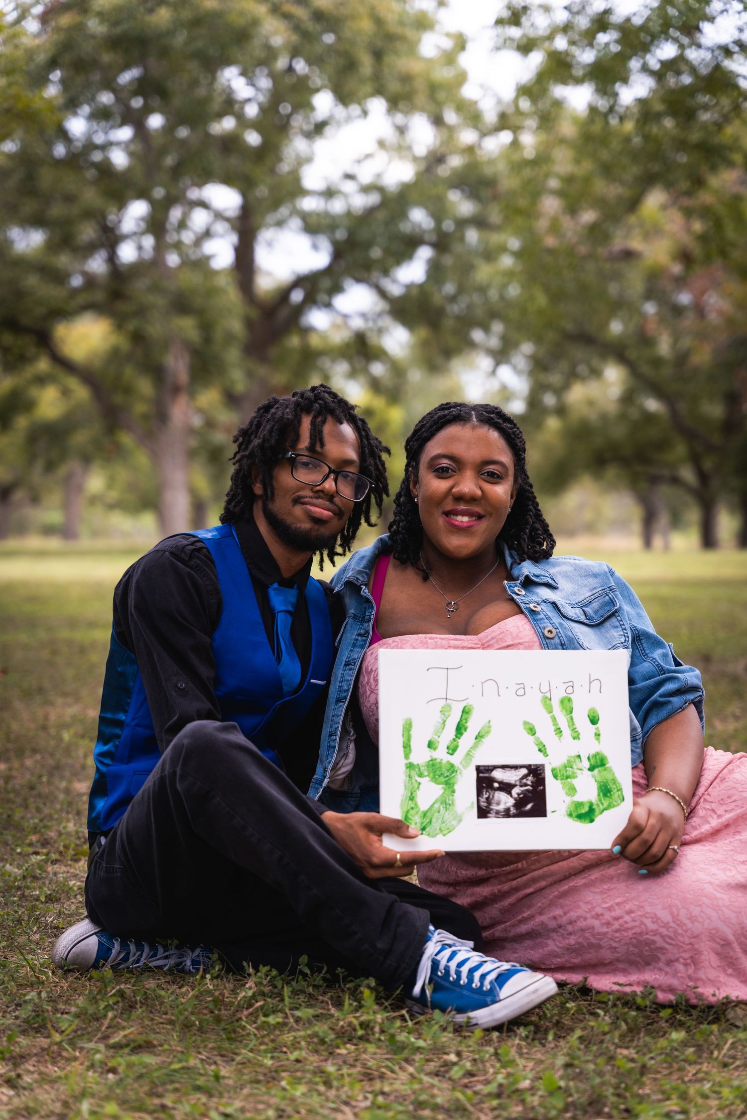A couple sits on grass holding a sign with green handprints and ultrasound photo during maternity shoot.