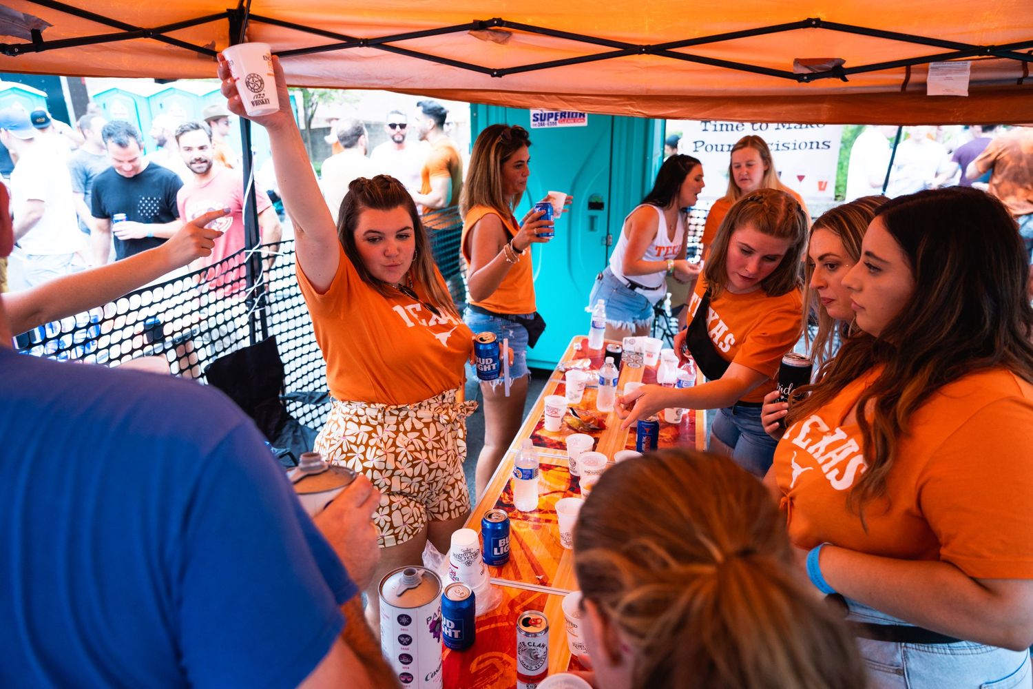 Group of people in orange shirts serving drinks at a festival booth under a colorful tent.
