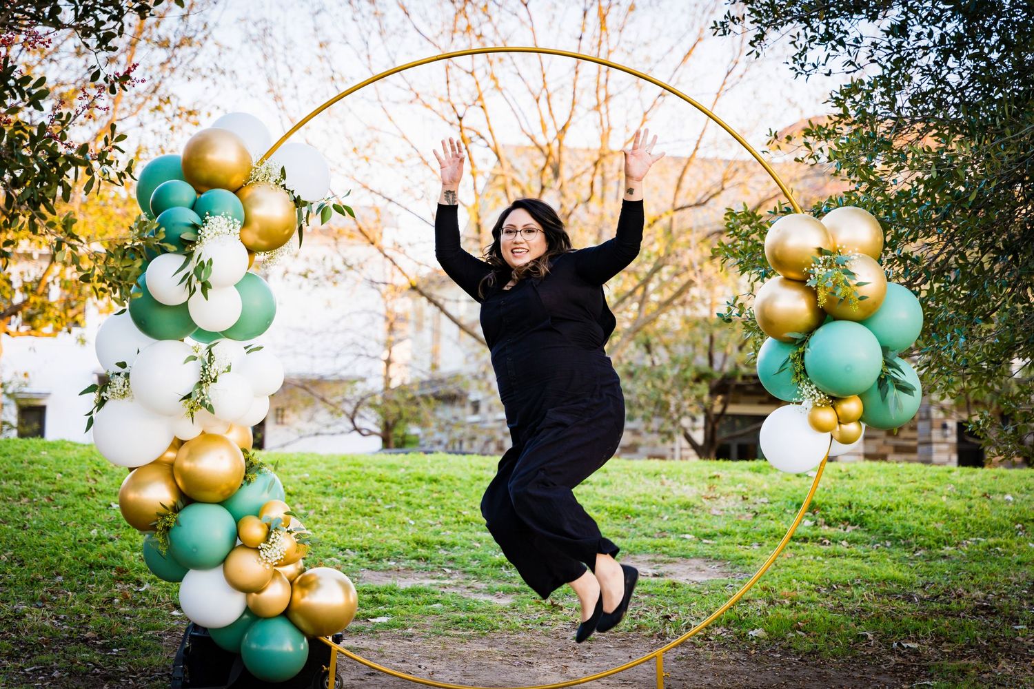 Person jumping joyfully in front of a decorative balloon arch with gold and mint green balloons outdoors.