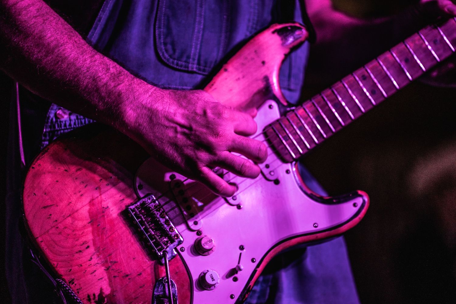 Close-up of hands playing pink-lit Stratocaster electric guitar during live performance.