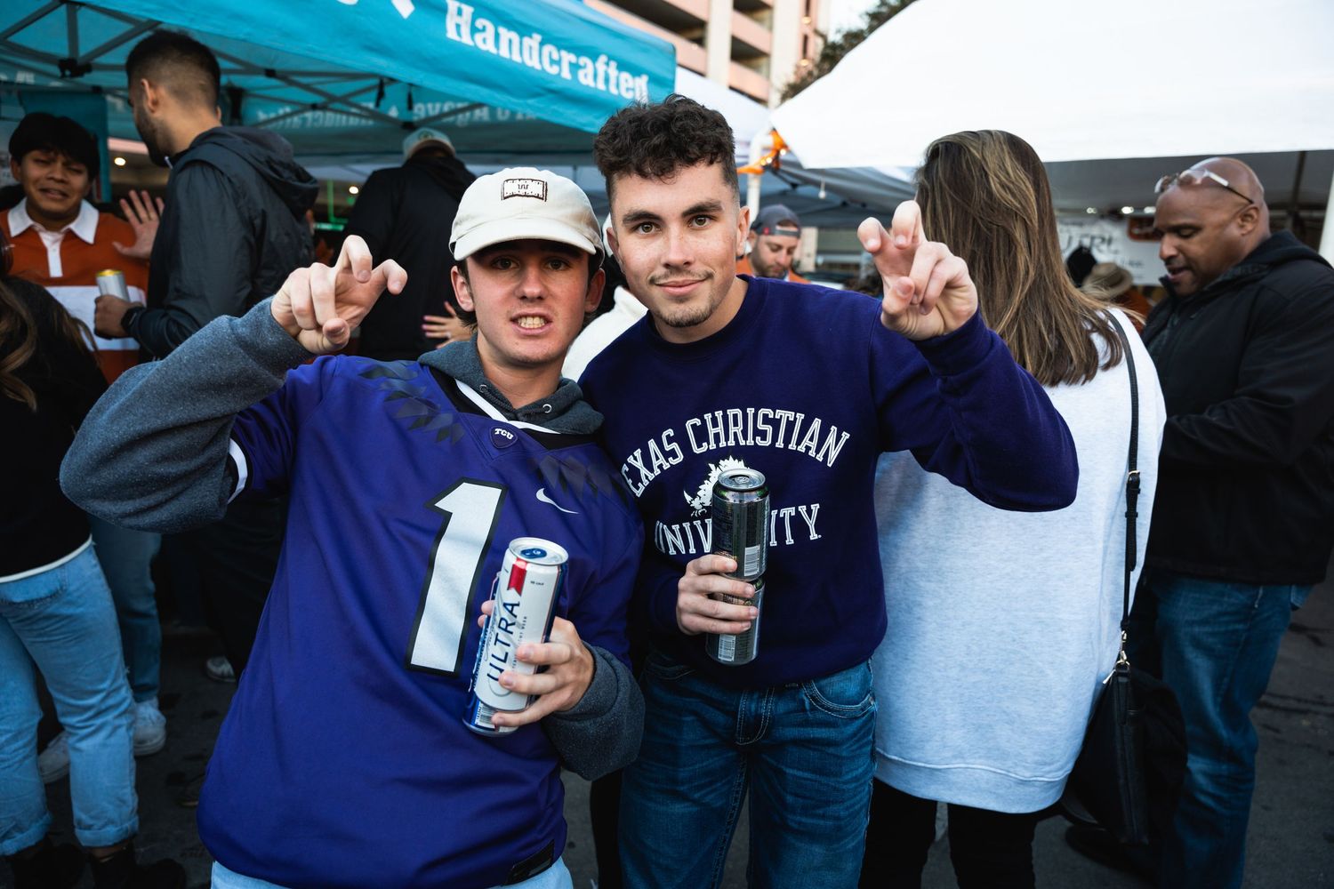 Two fans in college apparel celebrating at an outdoor event with beverages.