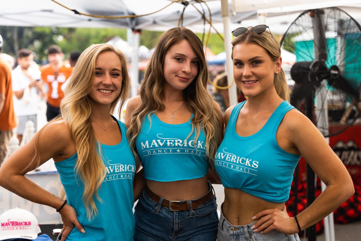 Three promotional staff members in matching blue tank tops pose at an event booth.