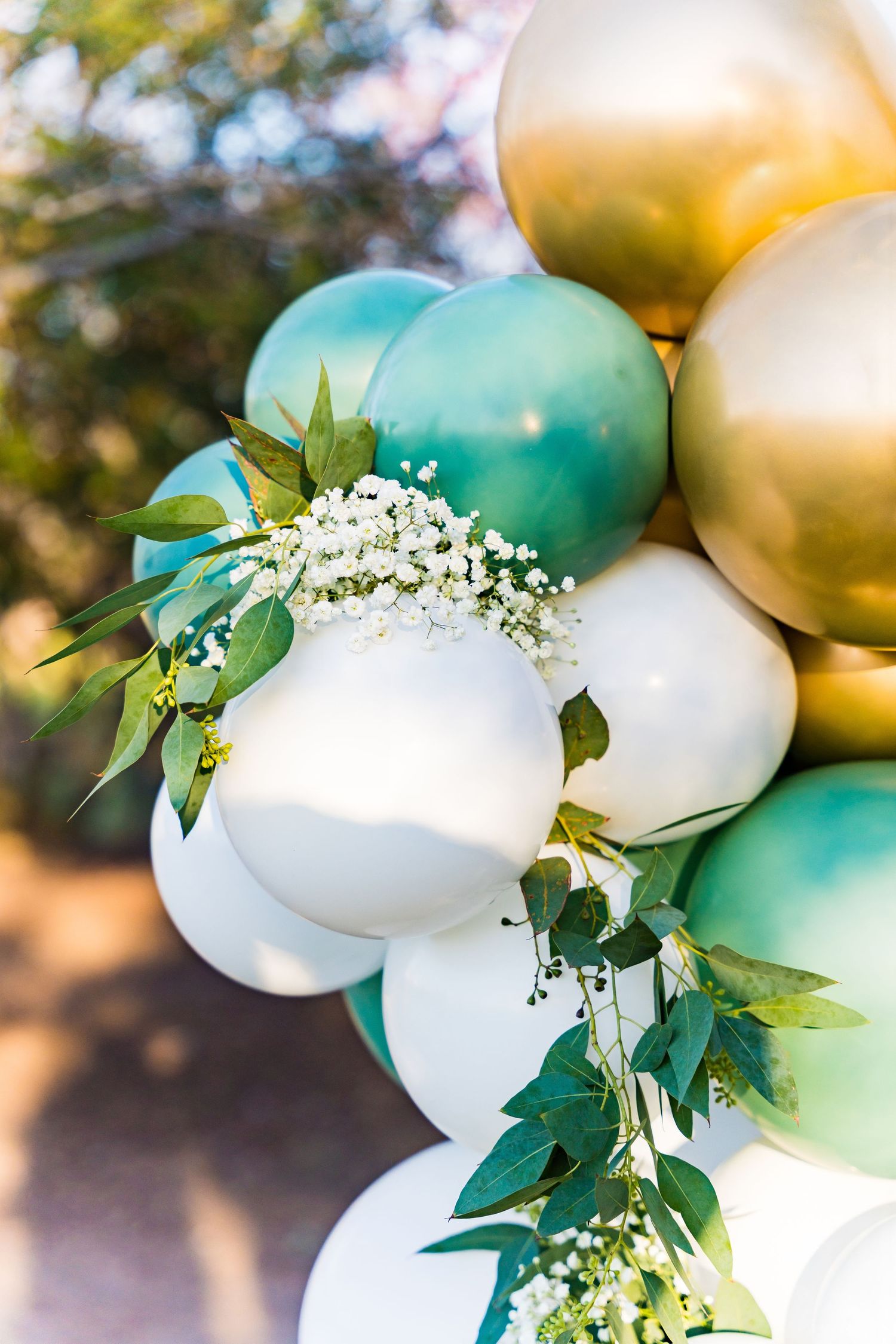 Elegant balloon arch decorated with gold, teal, and white balloons adorned with delicate white flowers and greenery.