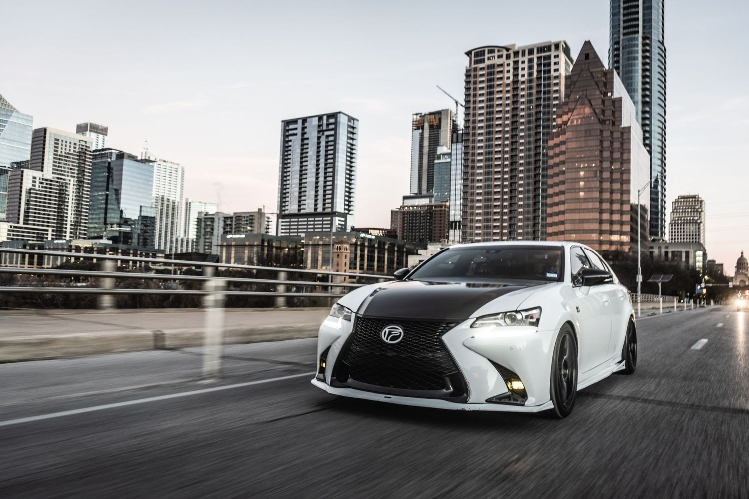 Customized white Lexus sedan driving on city street with downtown Austin skyline in background.