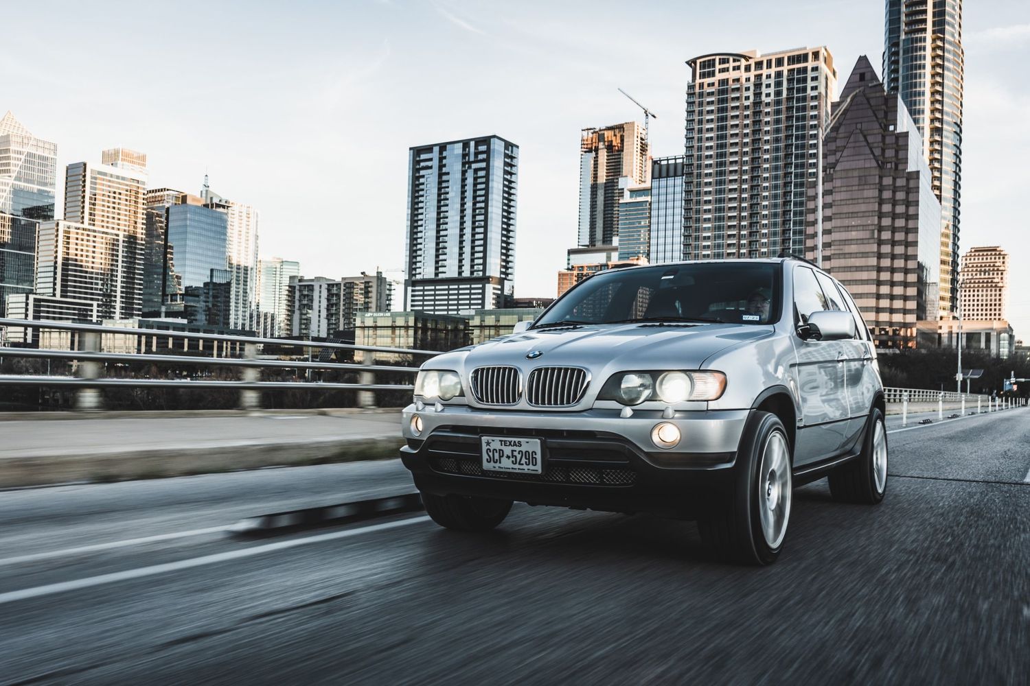 BMW X5 crossover speeds through downtown with modern skyscrapers forming dramatic urban backdrop.