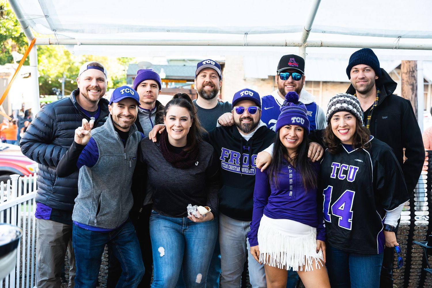 Group wearing purple sports fan gear posing together under a white tent at a tailgate party.