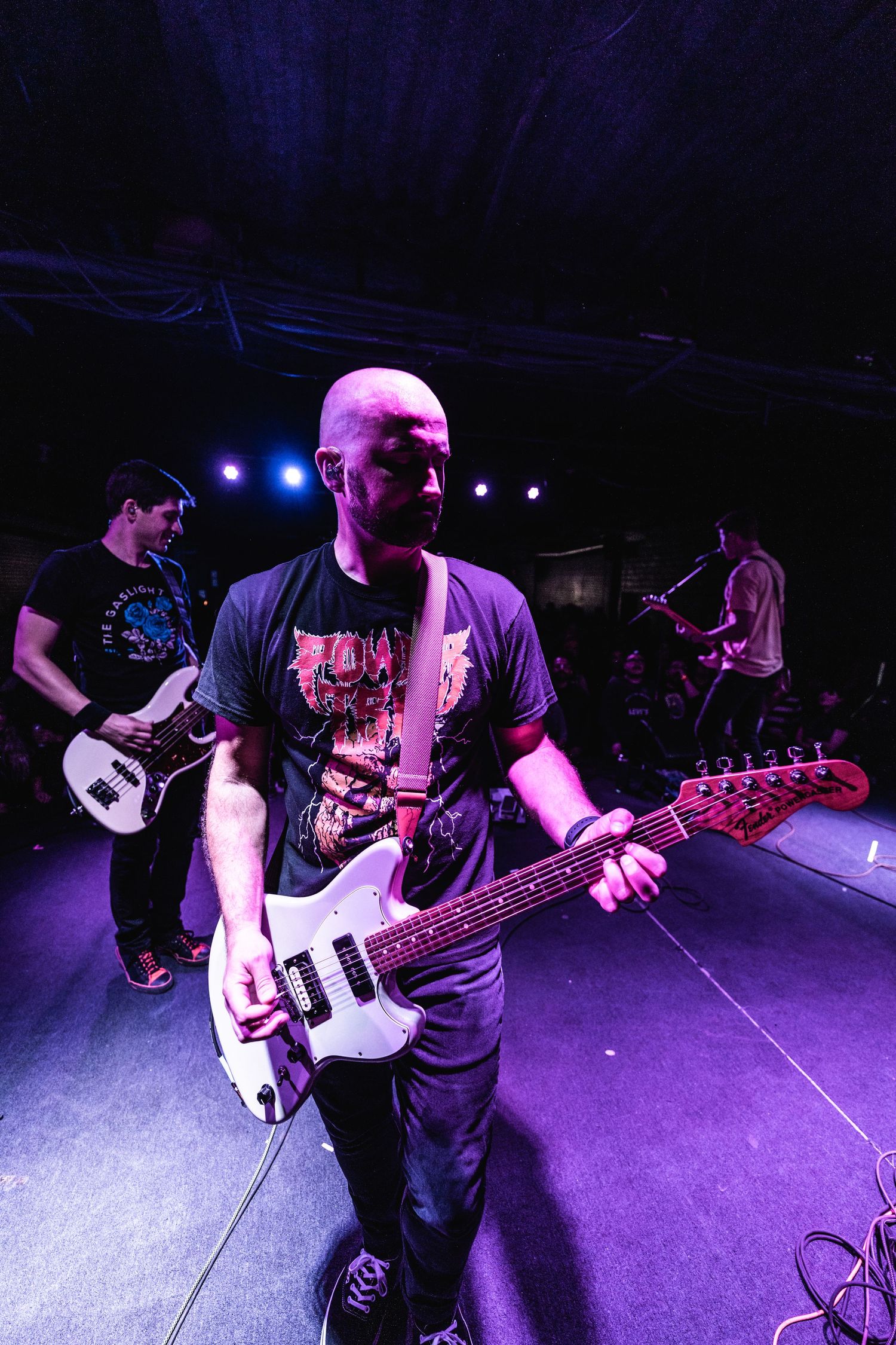 Guitarist performs with white electric guitar under purple stage lighting.