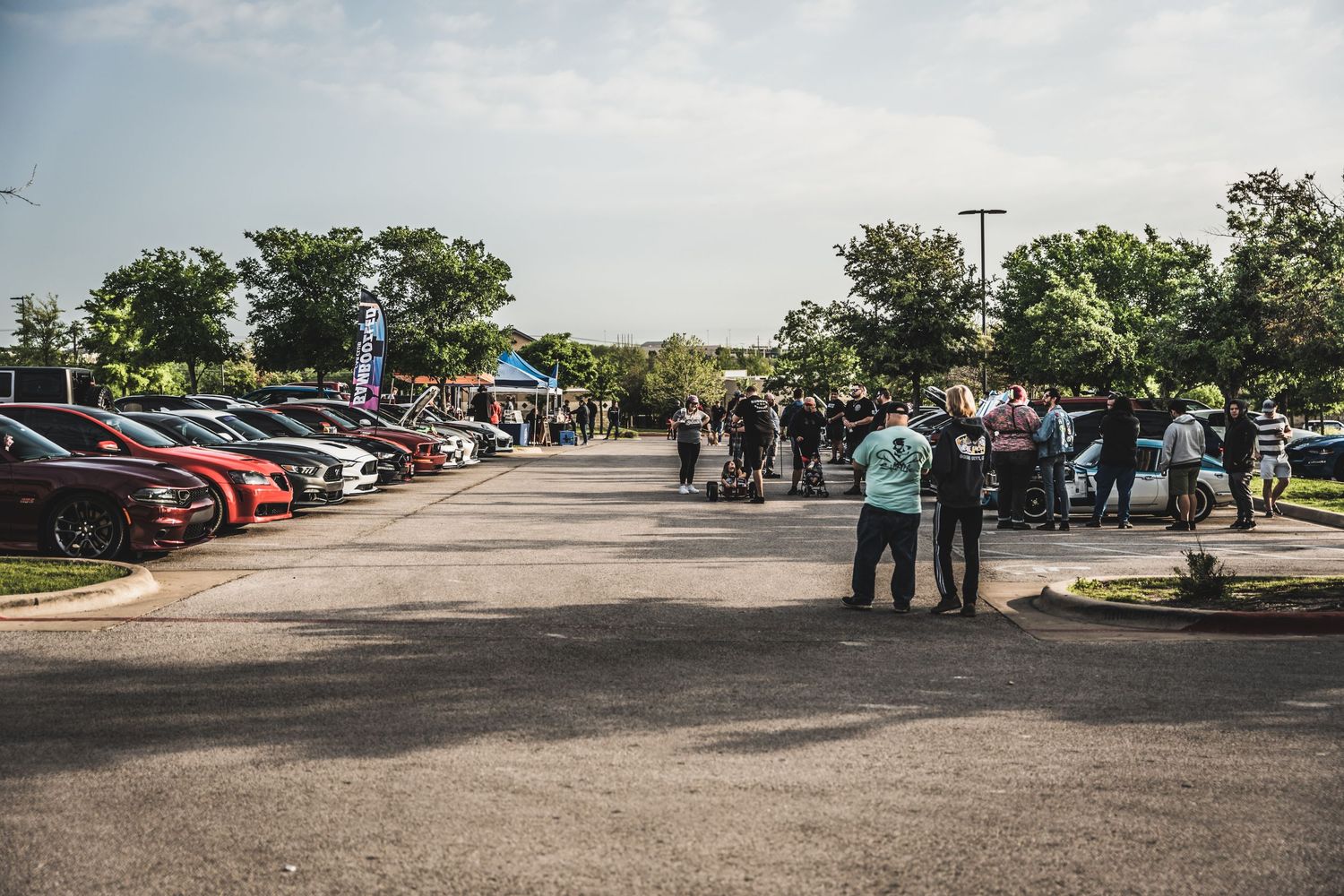 Wide view of a car show with numerous vehicles and attendees in a parking lot.