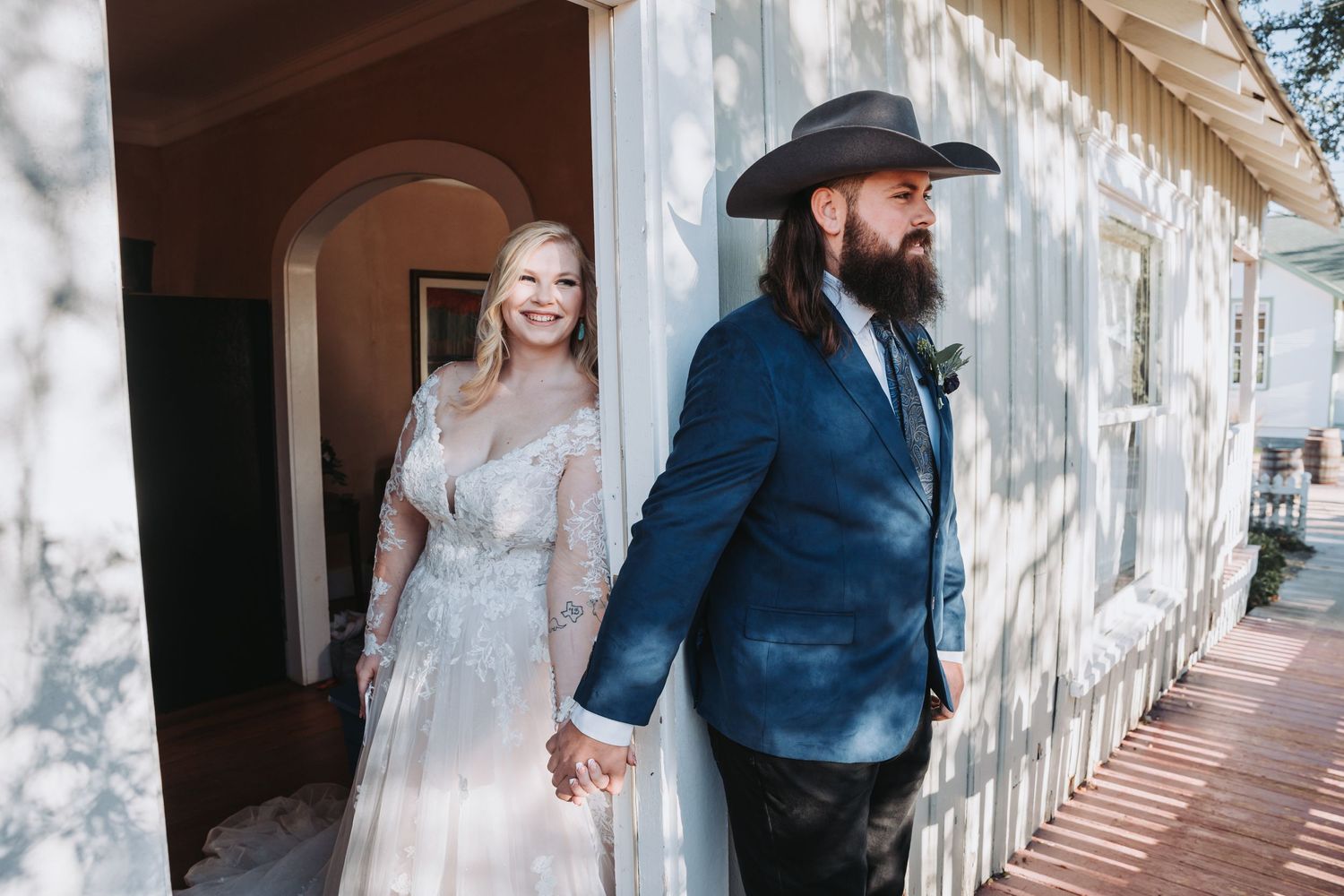 A couple in wedding attire stands on a rustic porch with one wearing a cowboy hat.