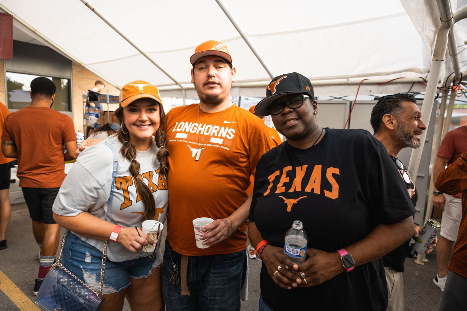Texas sports fans in orange and black team apparel gather under a white tent at a tailgating event.