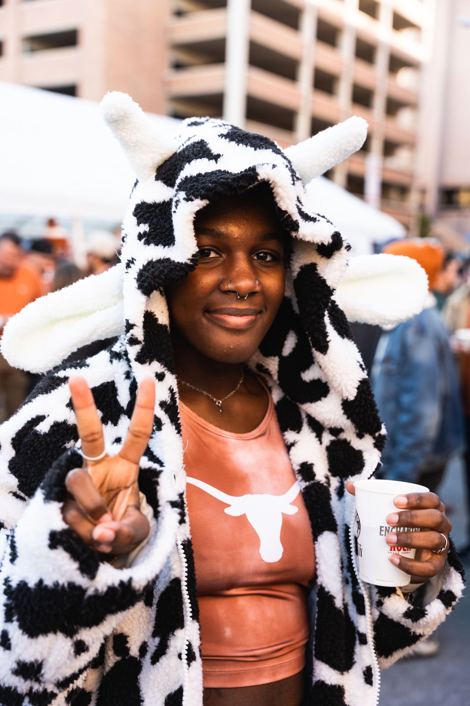 Person wearing a cow print hoodie and Texas shirt making peace sign at outdoor event.