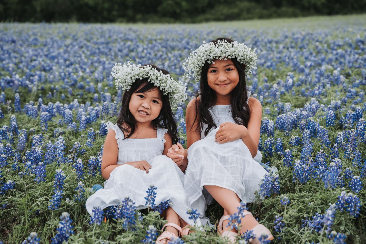 Two figures in white dresses sitting among blooming bluebonnets wearing floral crowns.