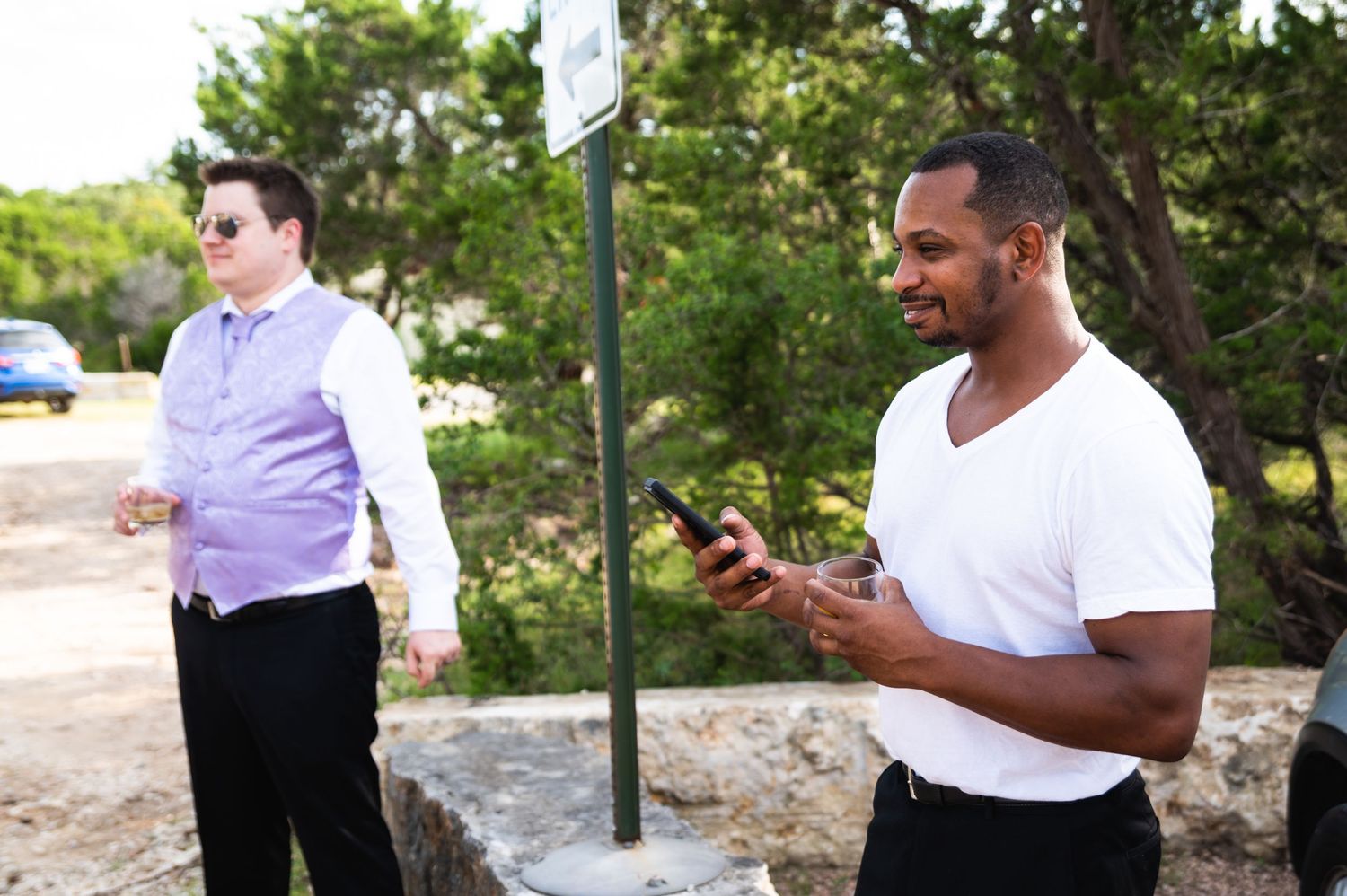 Two people standing outdoors, one wearing a purple shirt and another in white checking a mobile device.