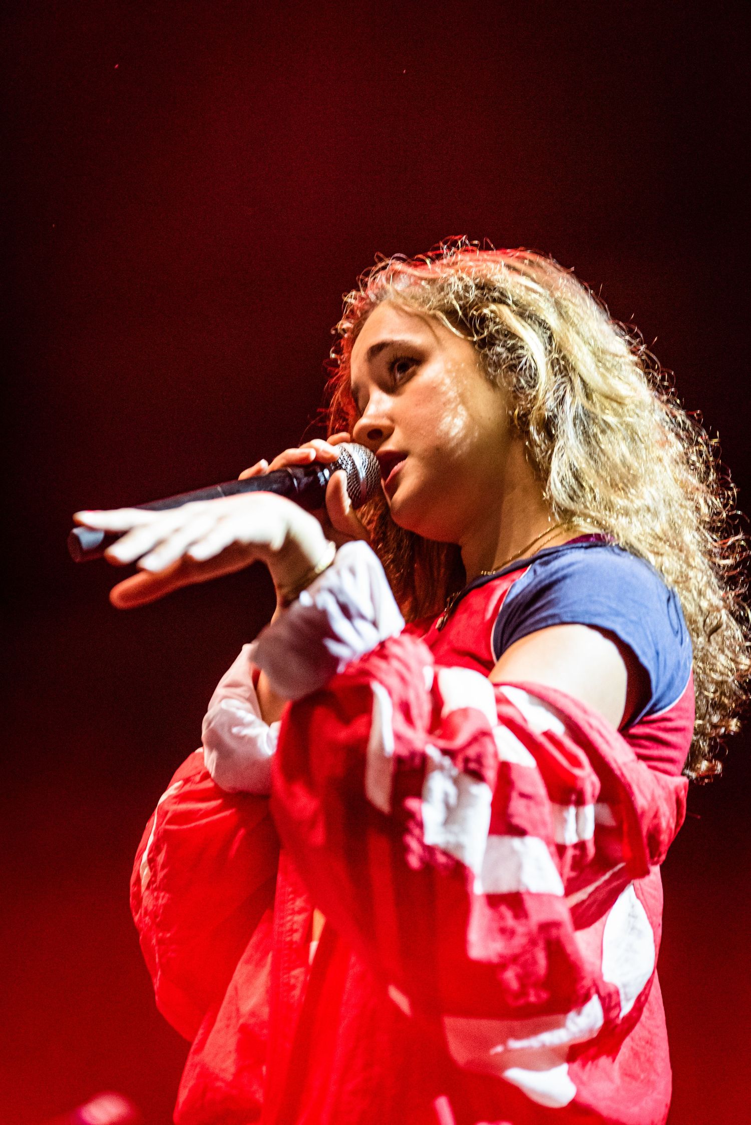 Stage performer in red jacket sings with dramatic gesture against red backdrop lighting.