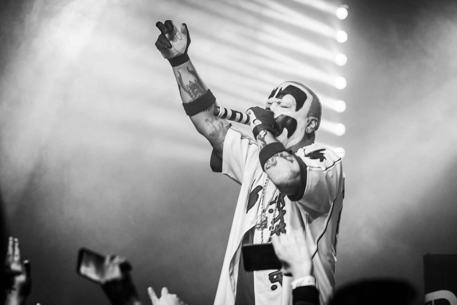 Performer in face paint and white costume raises arm dramatically during concert performance.