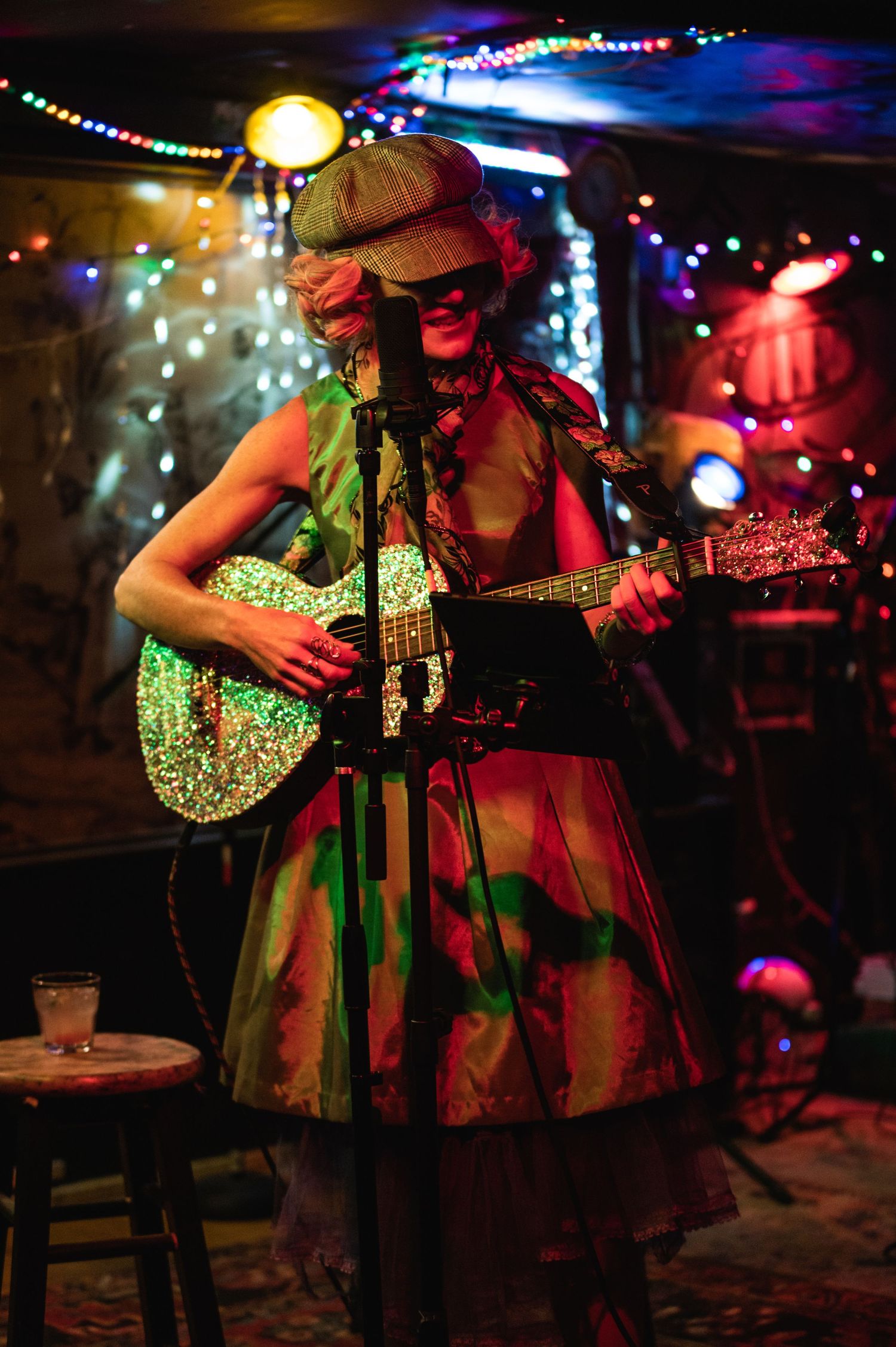 A musician in vintage dress plays guitar on stage with multicolored lights and decorations.