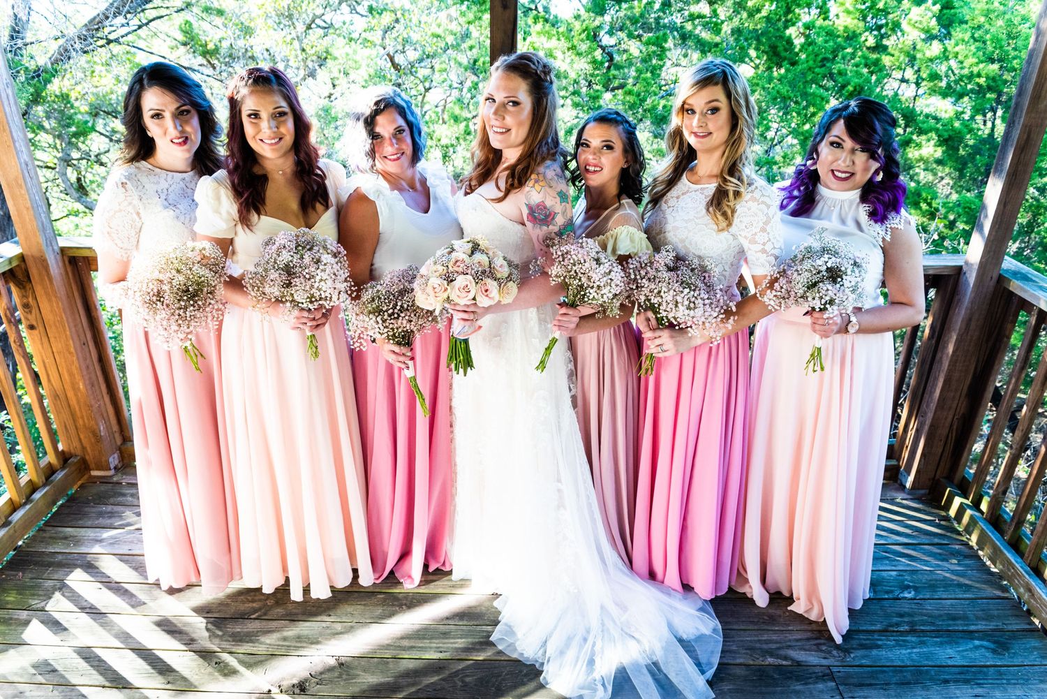 A wedding party poses on a wooden deck wearing pink bridesmaid dresses and holding delicate floral bouquets.